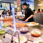 Interior scene of Yo! Sushi japanese conveyor belt restaurant at Westfield Stratford; a chef in black prepares dishes, food is moving along the conveyor belt, a shopping mall backdrop behind diners.