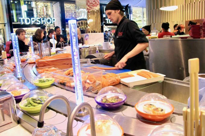 Interior scene of Yo! Sushi japanese conveyor belt restaurant at Westfield Stratford; a chef in black prepares dishes, food is moving along the conveyor belt, a shopping mall backdrop behind diners.