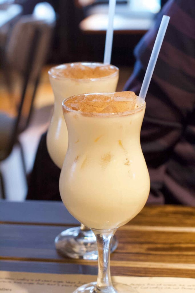 Pair of cream coloured Koko Kolada cocktails in tall stemmed curved hurricane glasses with clear plastic straws as served at Turtle Bay Caribbean style restaurant, Walthamstow branch East London. The floating ice cubes have been dusted with cinnamon. 