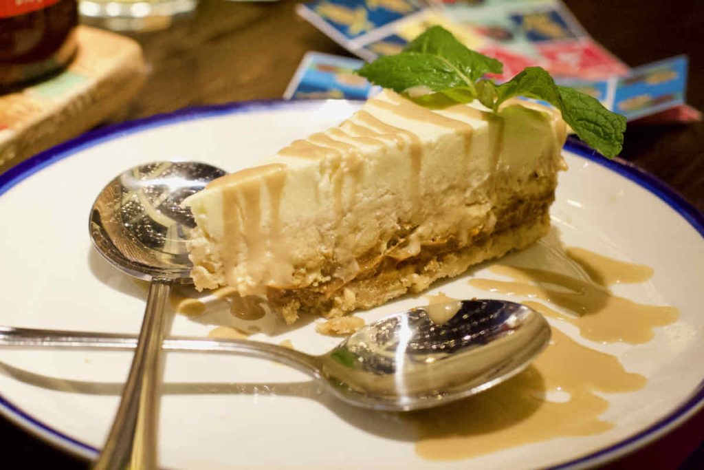 Slice of banana and toffee cheesecake garnished with mint sprig on a white plate with blue rim served with two spoons to share at Turtle Bay Caribbean style restaurant, Walthamstow branch East London.