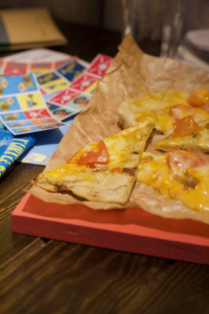 Triangles of toasted cheese and tomato flatbread on a sheet of brown paper and red board as served at Turtle Bay Caribbean style restaurant, Walthamstow branch East London. In the background are colourful stickers from the kids' entertainment pack. 