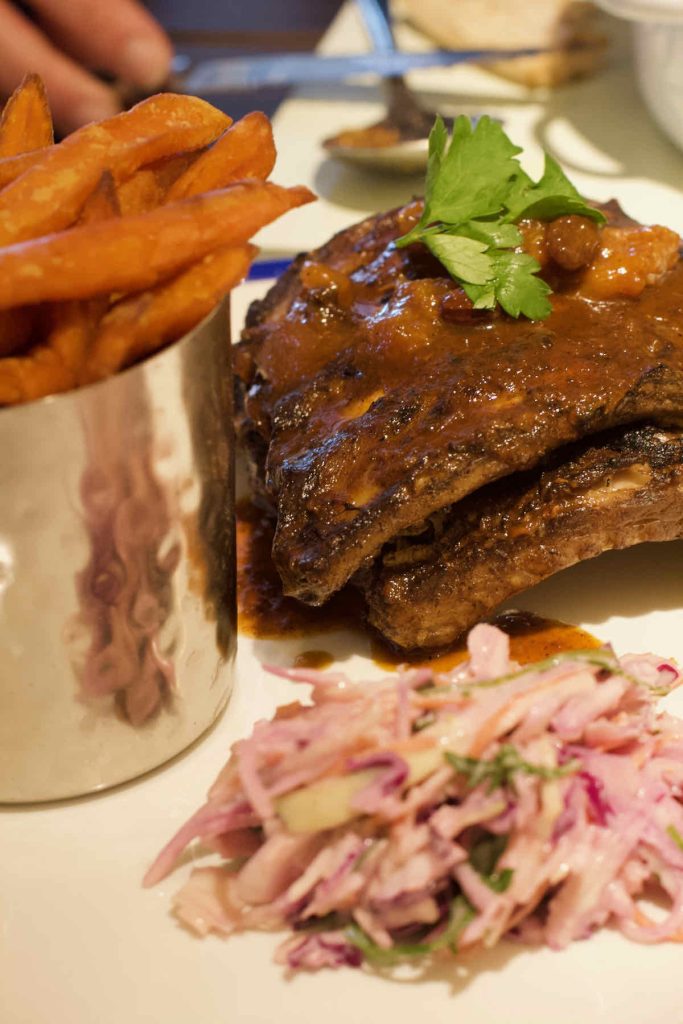 Jerk pork ribs covered in sauce served alongside sweet potato fries in a steel cylindrical side dish and some red onion coleslaw. From Turtle Bay Caribbean style restaurant, Walthamstow branch East London.