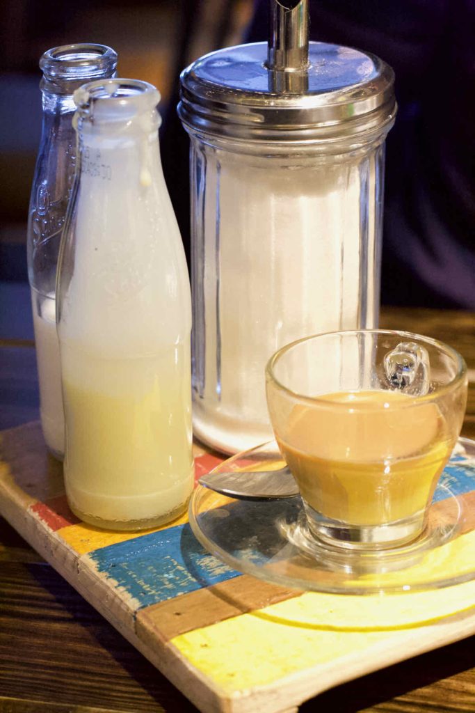 Tea in a small glass cup and saucer alongside glass bottles of both milk and condensed milk as served on a colourful painted table at Turtle Bay Caribbean style restaurant, Walthamstow branch East London. A giant sugar canister with aluminium spout stands in the background. 