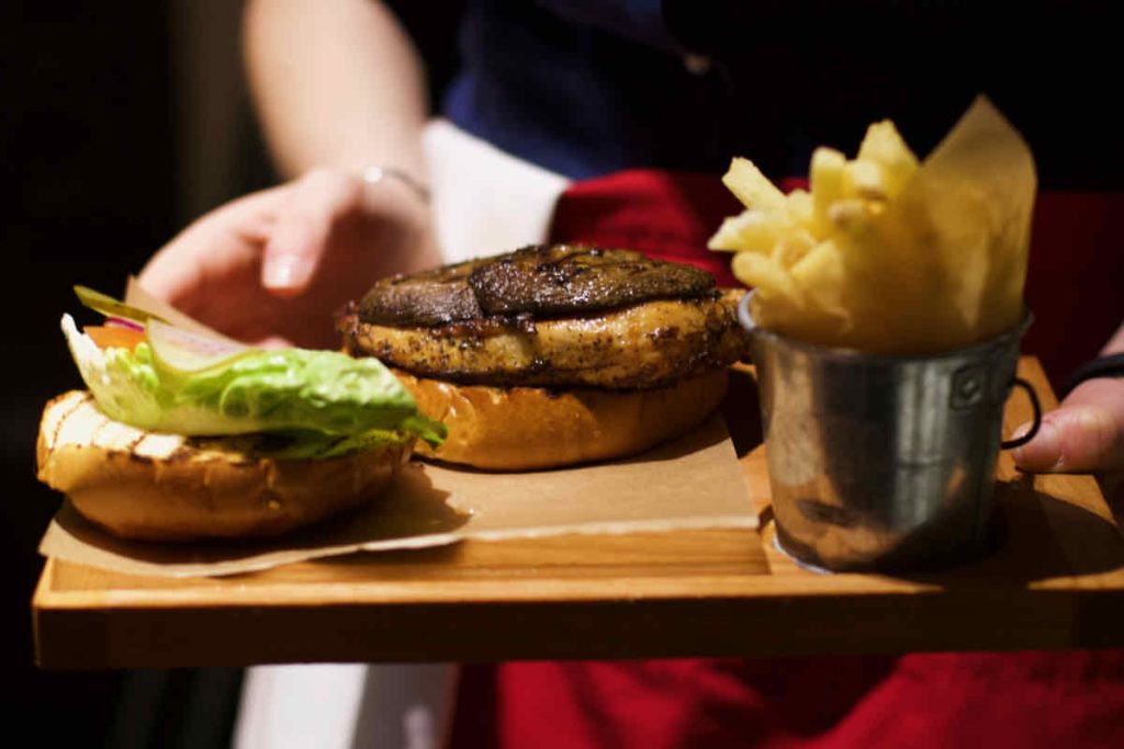 The hands of a server in dark top and red apron presents a blackened chicken burger with French fries upon a wooden board; as served at All Star Lanes bowling alley, Westfield Stratford, East London.