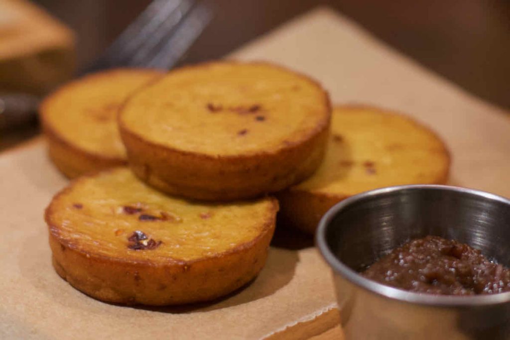 Golden "Hush Puppy" fritters, round cornmeal fried side dish item served on brown paper with a stainless steel pot of brown dip in the foreground; as served at All Star Lanes bowling alley, Westfield Stratford, East London.