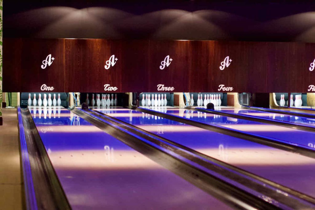 Interior view of the bowling alleys at All Star Lanes bowling alley, Westfield Stratford, East London. Lighting is pink and purple upon light wood bowling lanes with white bowling pins stood ready to play; lanes are marked with the branding "A*" and numbered one to four.