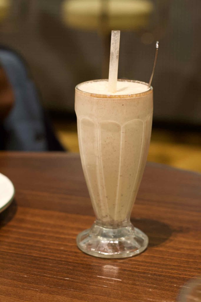 Light brown peanut butter milkshake with clear plastic straw in a tall ridge glass with foot, as served at All Star Lanes bowling alley, Westfield Stratford, East London. A tall spoon is sticking out of the glass.