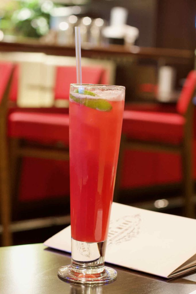 A tall slim glass of red lemonade mocktail with slice of lime, as served at All Star Lanes bowling alley, Westfield Stratford, East London. A bar menu is shown behind on the dark wooden table and red upholstered seating is shown as a soft focus background.