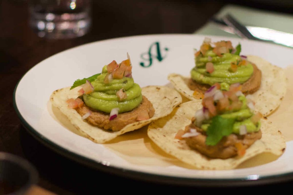 A trio of tostadas topped with swirls of refried beans, guacamole and chopped red onion and tomatoes lie on a white plate branded with the letter "A" upon a dark table. Some cutlery and a glass lie in soft focus behind. As served at All Star Lanes bowling alley, Westfield Stratford, East London