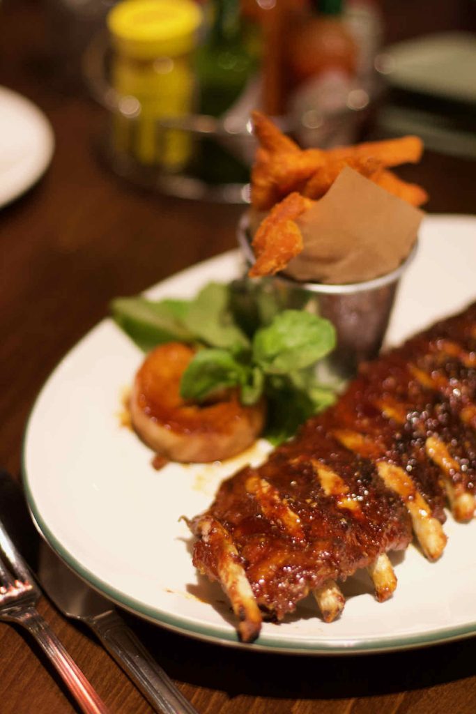 Main course of rack of ribs as served at All Star Lanes bowling alley, Westfield Stratford, East London; a golden "hush puppy" fritter, some green salad leaf garnish and a pot of orange sweet potato fries are also on the plate. Some condiments including yellow jar of Colman's mustard stand in soft focus behind.