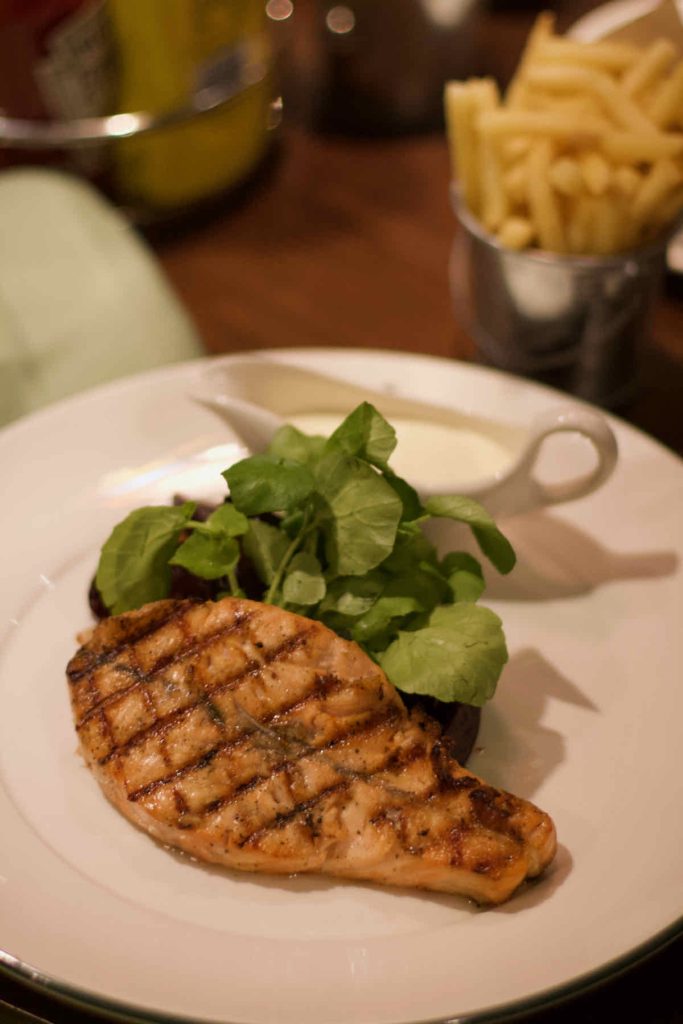 Salmon fillet as served at All Star Lanes bowling alley, Westfield Stratford, East London. Criss-cross grill marks have been made on the fish fillet, some green watercress leaves, a small sauce boat of white horseradish sauce and a steel pot of French fries lie in the background.