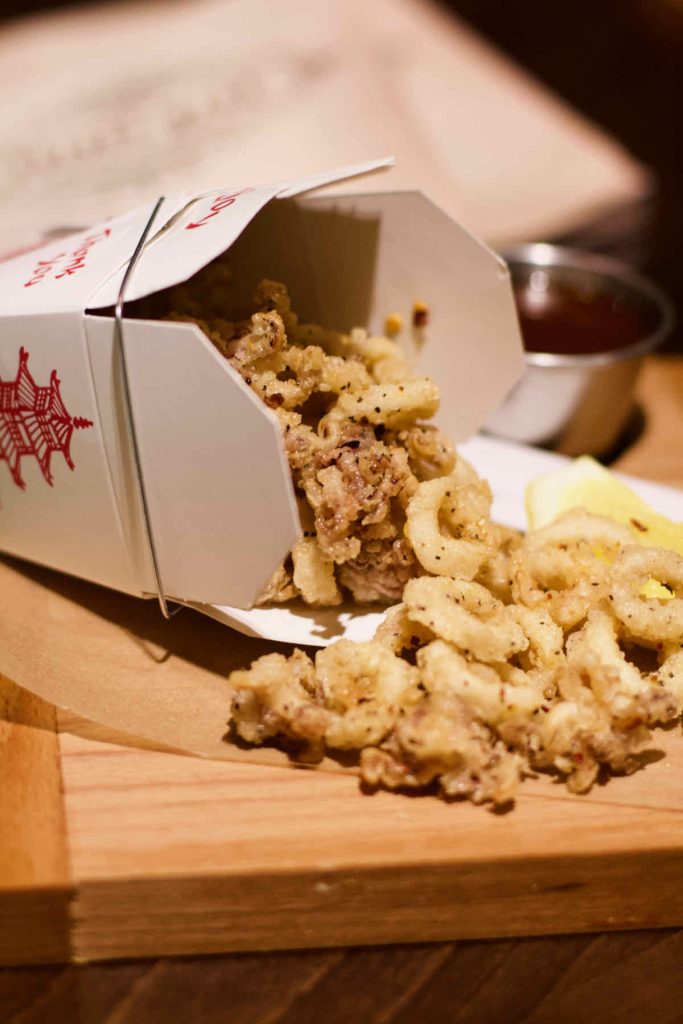 Salt and pepper squid as served at All Star Lanes bowling alley, Westfield Stratford, East London; Battered pieces of squid are tipped out of a cardboard American style Chinese take-out box onto brown paper upon a wooden serving board. A steel dish of dark sauce and a lemon wedge lie in soft focus behind.