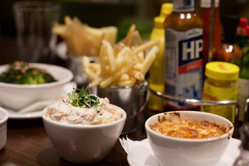 Various side dishes in small white or metal bowls as served at All Star Lanes bowling alley, Westfield Stratford, East London; from left to right, brocolli, coleslaw, French fries, macaroni cheese. Some bottles of condiments including HP brown sauce and Colman's mustard stand in the background.