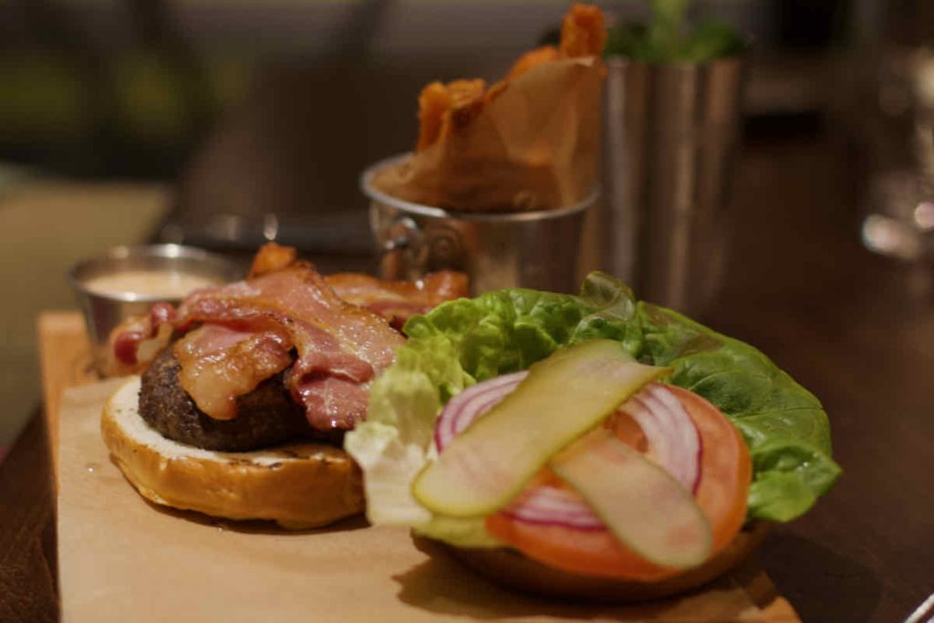 Classic bacon burger, presented open with patty and bacon on one half, salad and criss crossed gherkin on the other, as served at All Star Lanes bowling alley, Westfield Stratford, East London. Some metal serving pots stand in the background containing a mayo sauce and sweet potato fries.