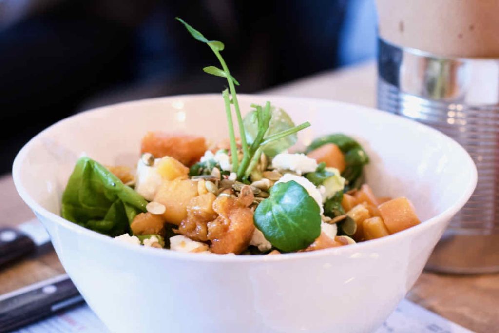 Butternut and feta salad, in a white ceramic bowl, as served at Cabana Brazilian barbecue restaurant at Westfield Stratford branch East London. In the background of the shot, a steel cylindrical tin with brown paper (containing chips) and two black wooden knife handles are shown.