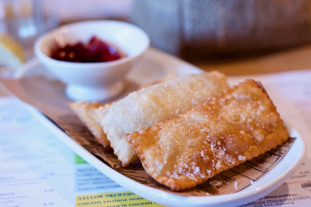 A trio of golden rectangular cheese pastries served on brown paper on an oval white dish with a smaller dish of red dip in soft focus behind. The pastry dish is upon the menu with colourful typography shown underneath; as served at Cabana Brazilian barbecue restaurant at Westfield Stratford branch East London.