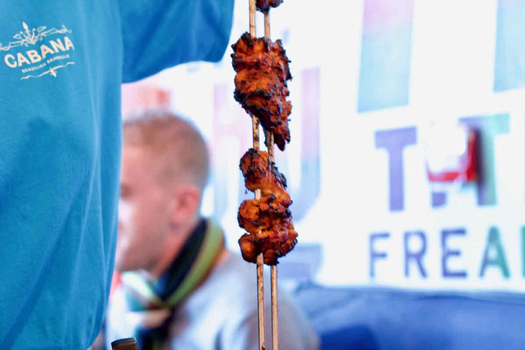 Vertical metal skewers of blackened barbecued chicken as served at Cabana Brazilian barbecue restaurant at Westfield Stratford branch East London. The server is partly seen in shot with a bright blue Cabana branded short-sleeved t-shirt. A man in a scarf is partly seen in soft focus behind with a wall displaying posters with large block letters of Portuguese words. The meat is red suggesting it is marinaded in spices.