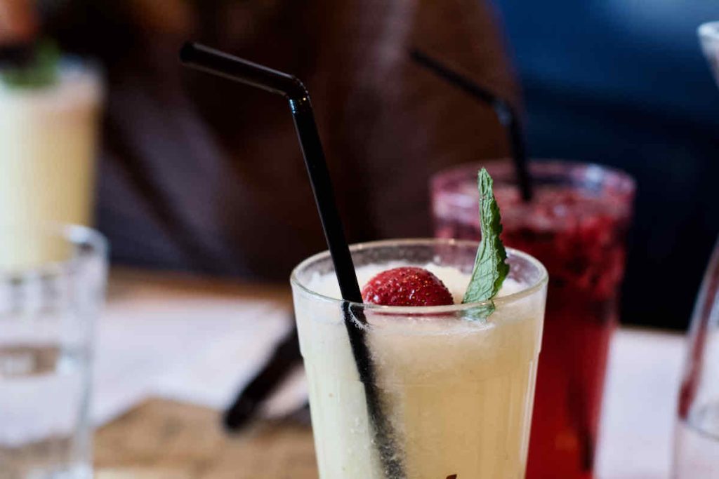 Pale yellow pineapple colada cocktail with black plastic straw, strawberry and mint garnish, as served at Cabana Brazilian barbecue restaurant at Westfield Stratford branch East London. A red drink is shown in soft focus behind.