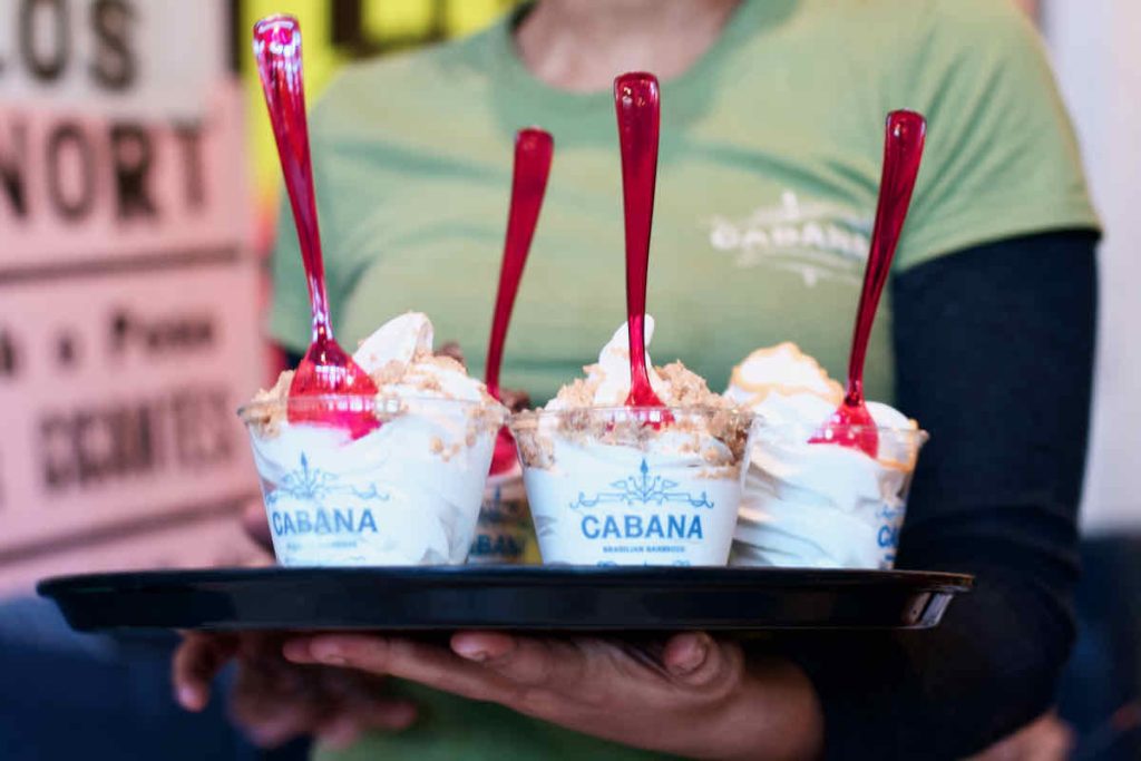 Clear plastic dishes of swirls of frozen yogurt with vertical pink plastic spoons sticking out of them, as served at Cabana Brazilian barbecue restaurant at Westfield Stratford branch East London. The server is partly in view holding the dishes on a black tray, their hands visible and flat under the tray. They are wearing a green short-sleeved Cabana t-shirt with a long-sleeved black t-shirt underneath. Poster signage from the restaurant interior is shown in soft focus to the side.