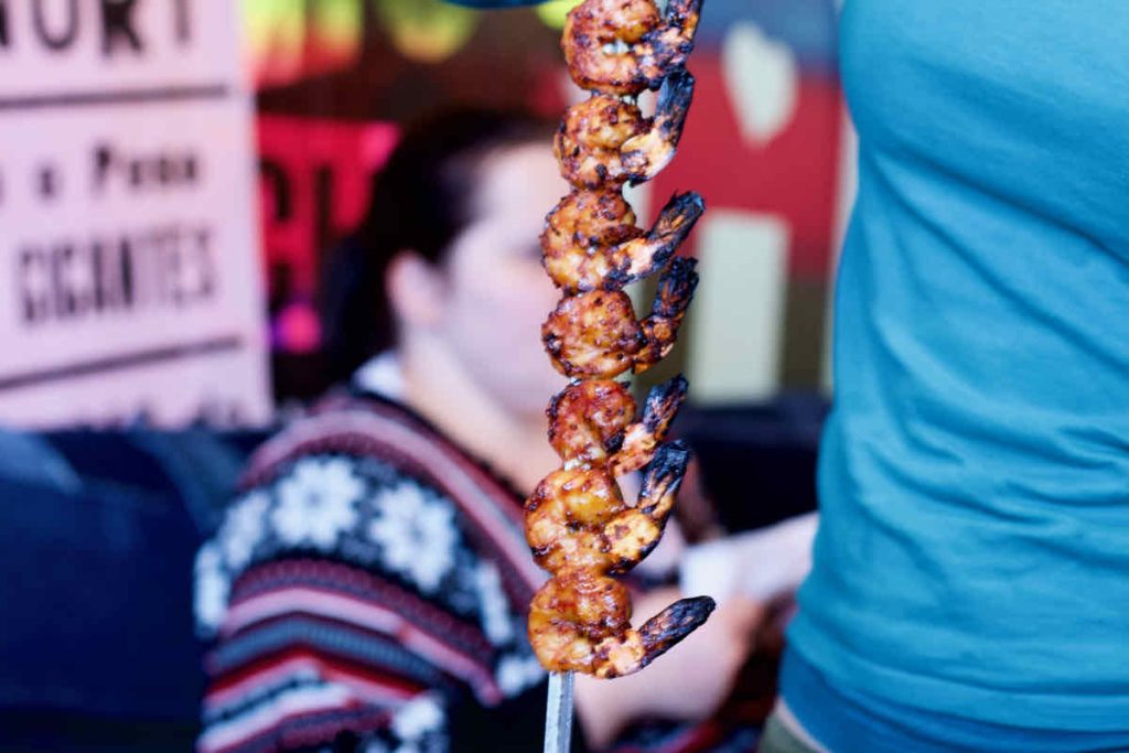 A metal skewer of eight barbecued king prawns on skewers as served at Cabana Brazilian barbecue restaurant at Westfield Stratford branch East London. The server is partly in shot with a turquoise t-shirt. In soft focus, a woman in patterned jumper is seated behind with colourful posters on the wall.