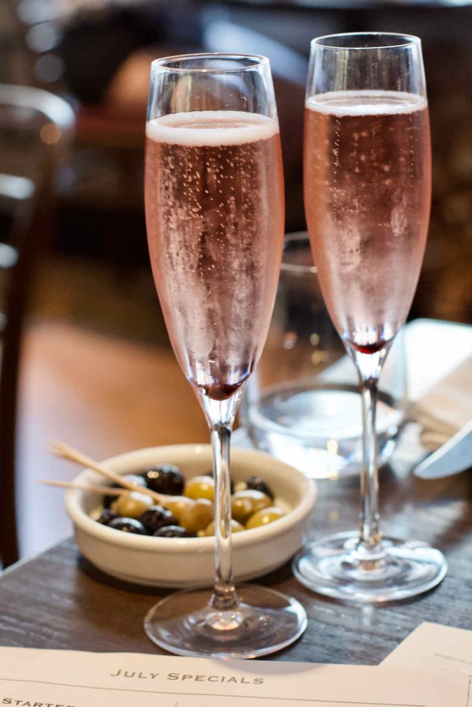 Two flute glasses of sparkling pink rosé wine, Kir Royale style, served on a dark wooden table at Cote Brasserie, Barbican Centre branch in the City of London. A small dish of green and black olives with cocktail sticks lies behind in soft focus.