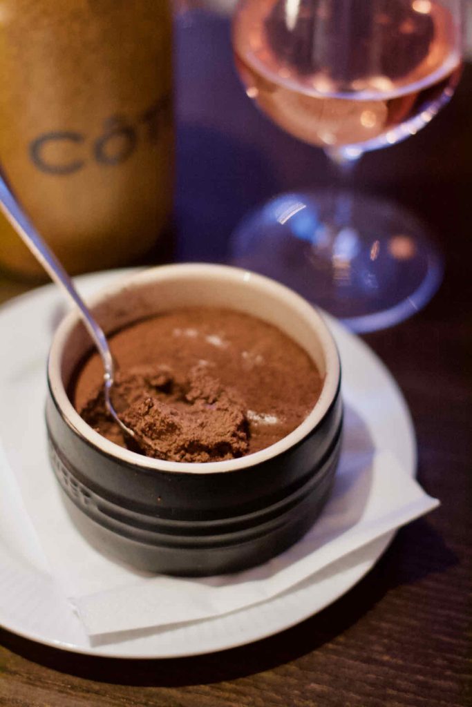 A spoon dipped into a brown Le Creuset branded ramekin filled with milk chocolate mousse; as served at Côte Brasserie, Barbican Centre branch in the City of London. A Côte branded water carafe and glass of rosé wine lie in soft focus behind.