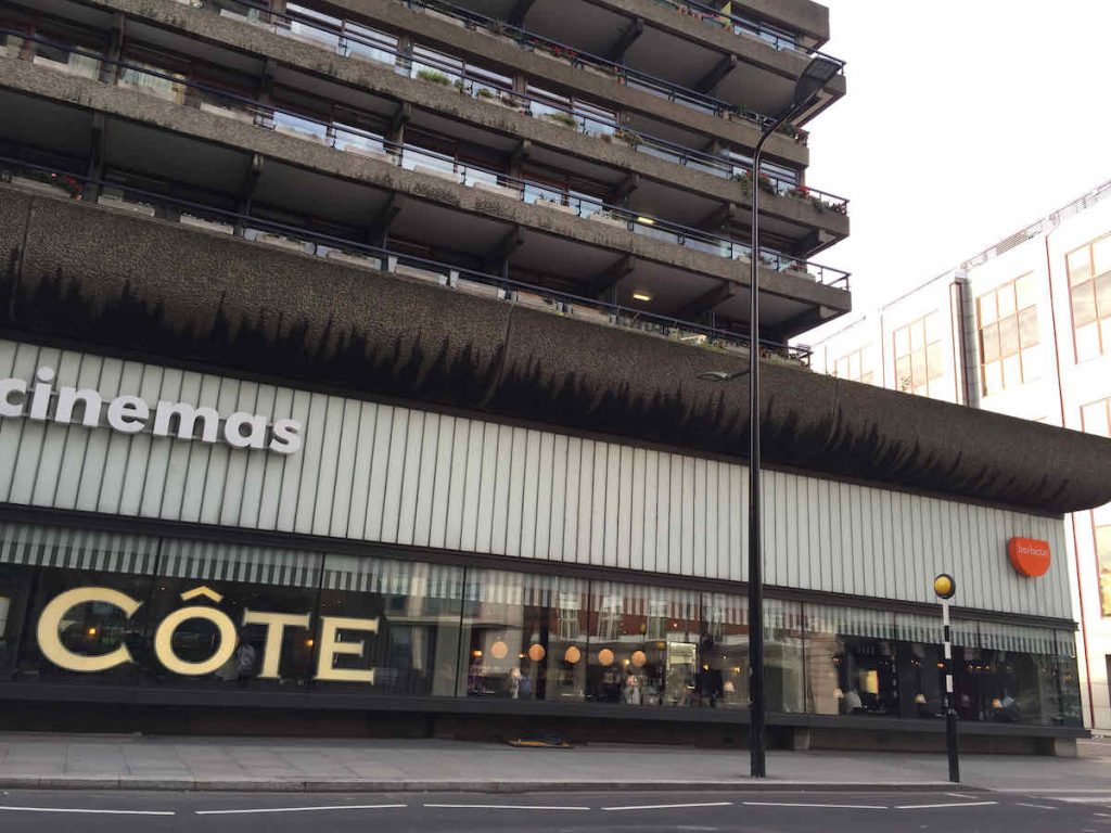 Côte Brasserie exterior view of French restaurant branch in the Barbican Centre, City of London. Several storeys of Brutalist flats typical of the Barbican Centre are seen above, a zebra pedestrian crossing with zig zag road markings is seen in front. The large windows have a giant gold coloured sign saying Côte. 