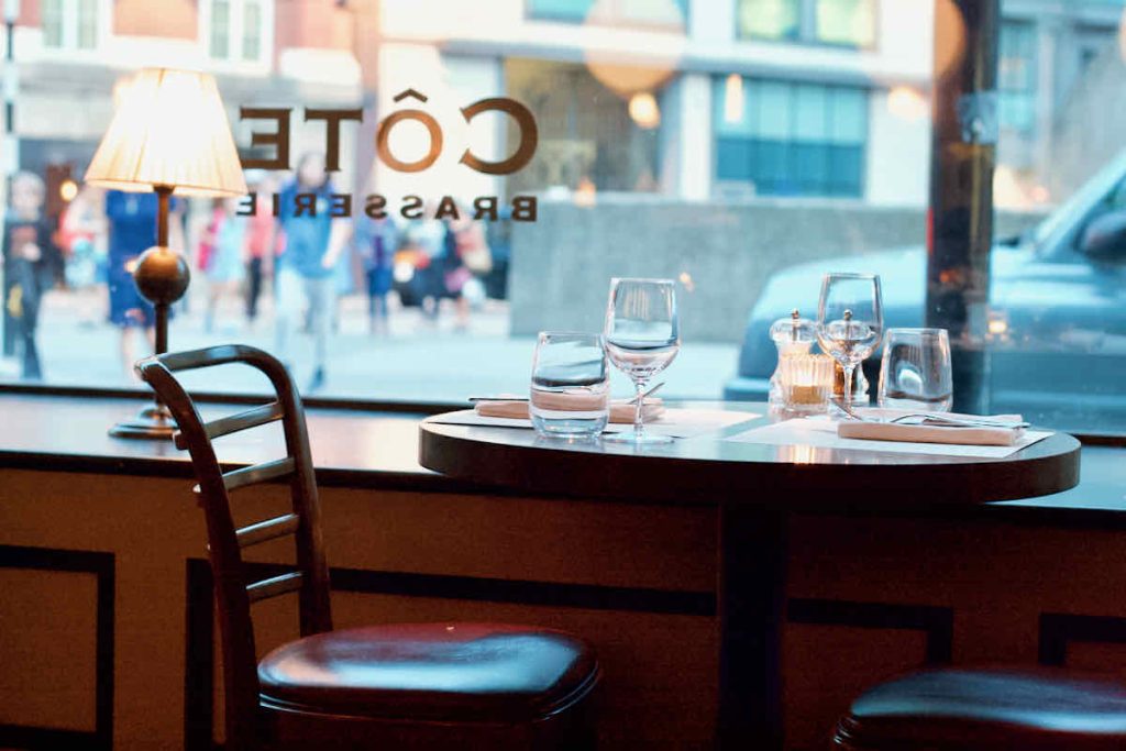 Côte Brasserie interior shot of window table with curved wooden chair, a round bistro table laid for two with glassware, candle, napkins and cutlery; the street outside with people is shown in soft focus. A table lamp with a white shade is lit on the dark wooden window ledge.