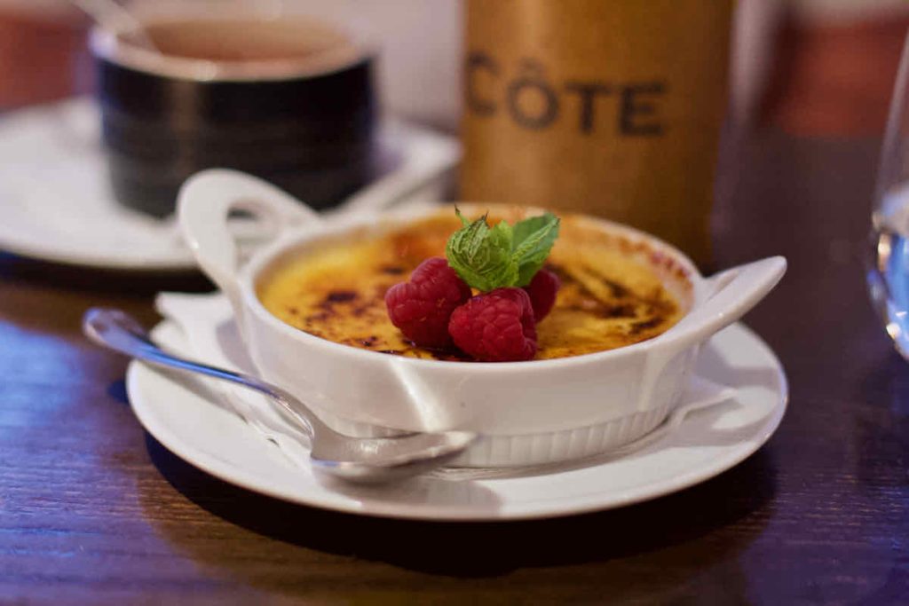 Lemon crème brûlée with mint and raspberry garnish, as served in a small white dessert oven dish with a spoon on a white side plate at Côte Brasserie, Barbican Centre branch in the City of London. A dark brown ramekin and Côte branded water carafe lies in soft focus behind on the dark wood table. 