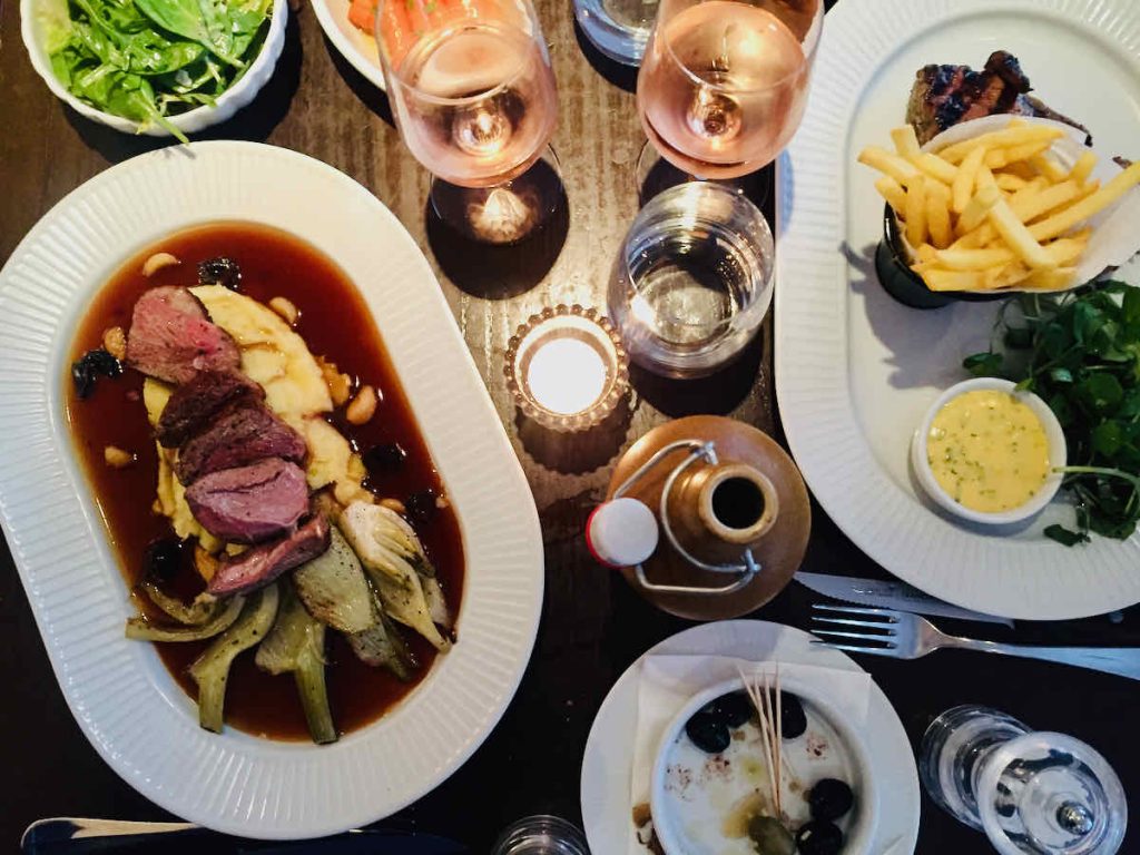 Overhead view of meal in French restaurant; rump of lamb with mashed potatoes, fennel and gravy, steak frites with Béarnaise sauce and watercress, both served on ridged oval-shaped white plates at Côte Brasserie, Barbican Centre branch in the City of London. Various glasses of water, rosé wine lie around the plates on the candle-lit dark wood table. 