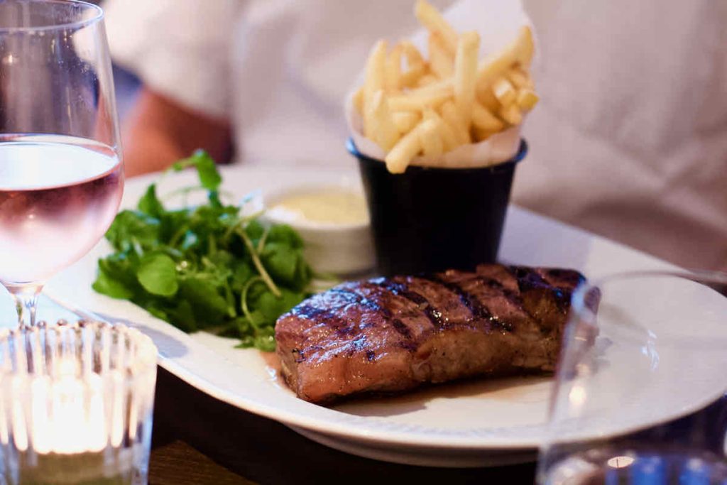 Medium rare steak, with side serving of green watercress and black pot of French fries styled in a white paper cone, as served on an oval white plate at Côte Brasserie, Barbican Centre branch in the City of London. In soft focus around the dish lies a candle in a tealight holder, a glass of rosé wine and a small dish of Béarnaise sauce. The backdrop is that of a man in a white short-sleeved shirt.