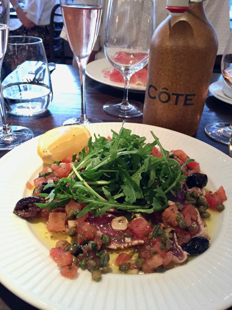 Tuna carpaccio with rocket, tomato, olives and capers and wedge of lemon, as served on a ridged white plate at Côte Brasserie, Barbican Centre branch in the City of London. A brown ceramic water carafe with Côte branding lies behind. Various glassware stands around the plate.