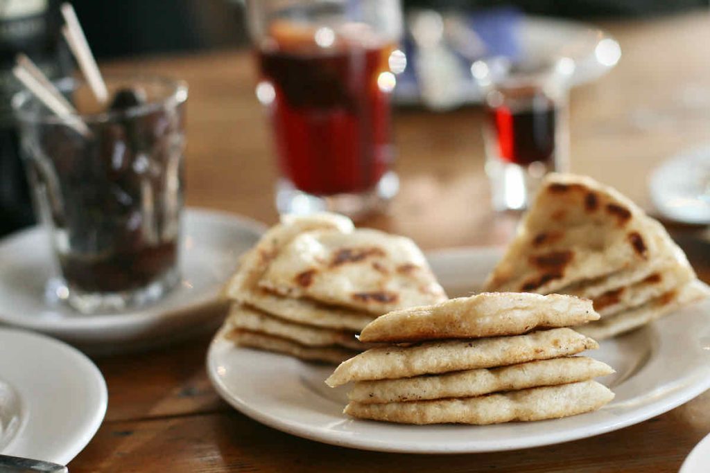 Triangles of flatbread piled on a white plate, glasses of black olives with cocktail sticks and red wine lie in soft focus in the background. As served at The Real Greek restaurant at Westfield Stratford, East London.