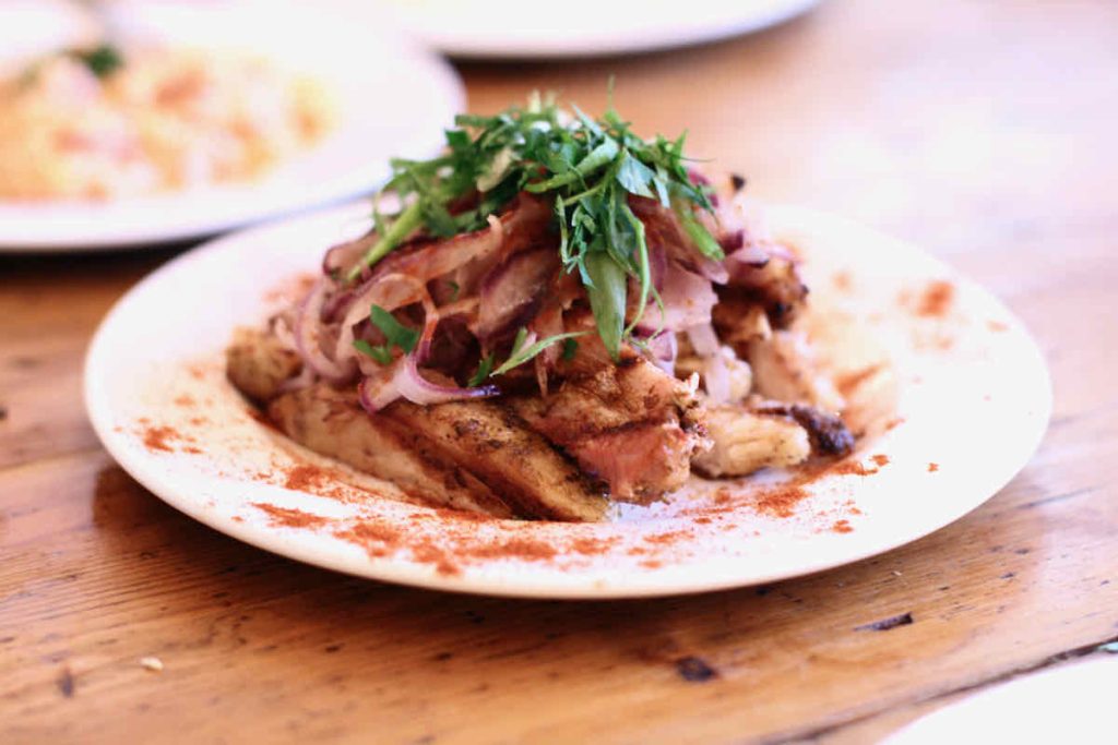 A pile of sliced pork belly garnished with red onion slices and green herbs upon a white plate dusted with red paprika, upon a vintage wooden table; as served at The Real Greek restaurant at Westfield Stratford, East London.