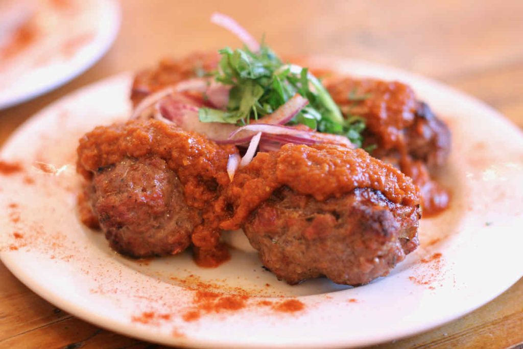 A white plate of Greek meatballs topped with bright red tomato sauce, slithers of red onion and chopped green herbs as served at The Real Greek restaurant at Westfield Stratford, East London.