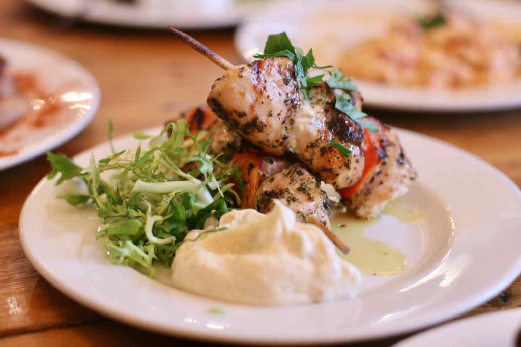 Plate of two small wooden skewers loaded with grilled chicken pieces upon a white plate with spiky salad leaves and large blob of mayonnaise, as served at The Real Greek restaurant at Westfield Stratford, East London.