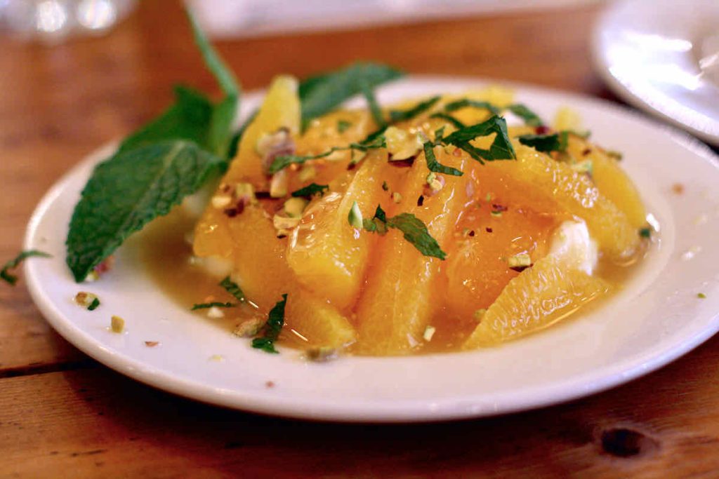 Bright segments of orange citrus flesh, skins removed topped with chopped mint and pistachio nuts and honey, upon a white plate as served at The Real Greek restaurant at Westfield Stratford, East London.