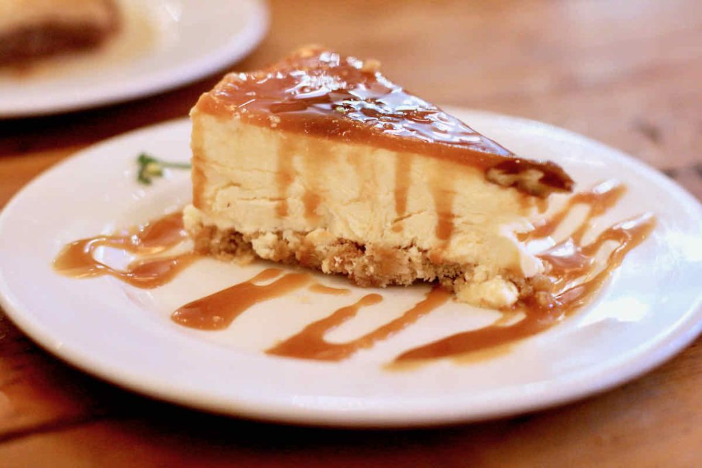 A large triangular slice of caramel and pecan cheesecake on a white plate zig zagged with caramel sauce. As served at The Real Greek restaurant at Westfield Stratford, East London.