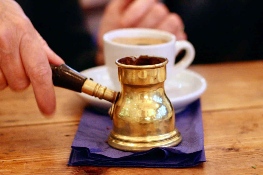 At a vintage wooden table, a small brass coffee pot is held by its dark wooden handle upon a pile of dark blue paper napkins. A white coffee cup and saucer, with someone else's hand lie in soft focus behind. As served at The Real Greek restaurant at Westfield Stratford, East London.