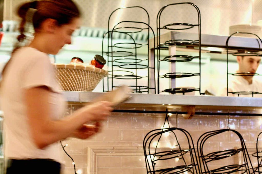 Interior shot of The Real Greek restaurant at Westfield Stratford, East London; a waitress in white short-sleeved t-shirt dashes past a stainless counter stacked with black metal serving stands, a chef stands in soft focus behind these. 