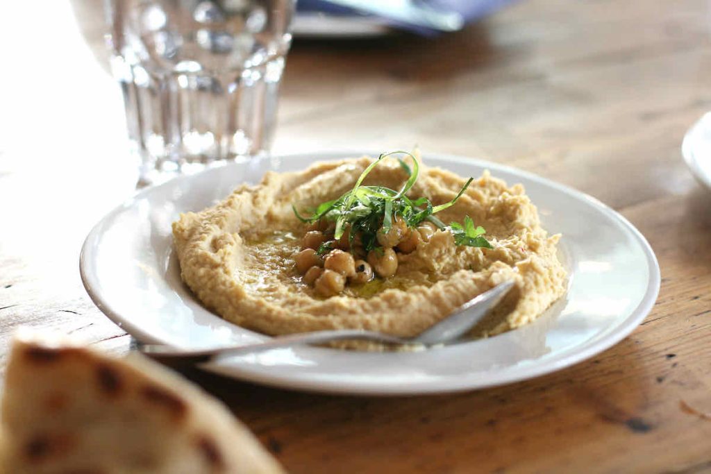 A plate of hummus on a white plate topped with chickpeas, herbs and glistening with olive oil; a spoon lies on the plate upon the vintage wooden table and in background soft focus lies a glass tumbler.