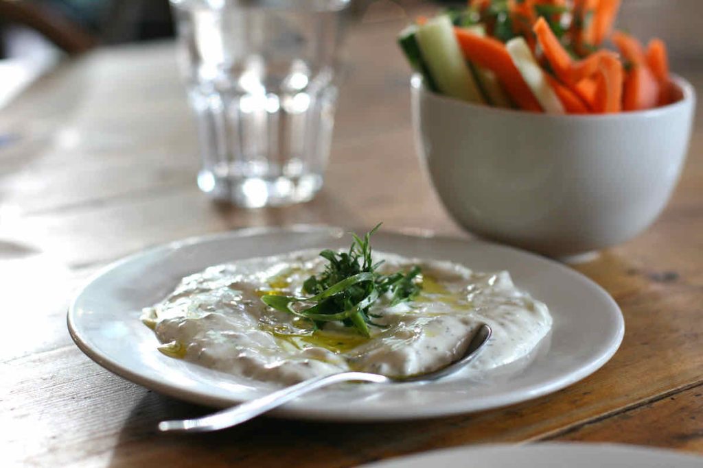 A plate of tzatziki cucumber and yogurt dip garnished with herbs on a white plate with a serving spoon; in the background in soft focus lies a glass tumbler and a white ceramic bowl filled with cucumber, carrot and pepper batons. All lie on a vintage wooden table. As served at The Real Greek restaurant at Westfield Stratford, East London.