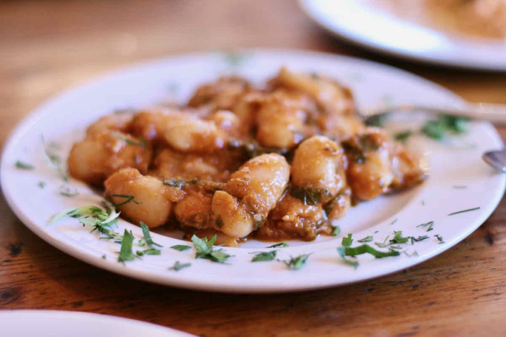 A white plate of gigandas-plaki or "gigantic beans" scattered with chopped herbs, as served at The Real Greek restaurant at Westfield Stratford, East London.