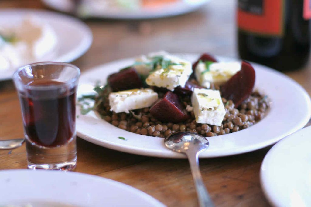 A white plate of puy lentil salad topped with large pieces of feta cheese and beetroot as served at The Real Greek restaurant at Westfield Stratford, East London. Adjacent is a shot glass of red sweet wine. A serving spoon, handle facing towards the foreground, lies face down on the plate. In the background of the vintage wooden table, a wine bottle with red label and a white plate with another side dish are shown in soft focus. 