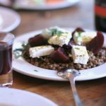 A white plate of puy lentil salad topped with large pieces of feta cheese and beetroot as served at The Real Greek restaurant at Westfield Stratford, East London. Adjacent is a shot glass of red sweet wine. A serving spoon, handle facing towards the foreground, lies face down on the plate. In the background of the vintage wooden table, a wine bottle with red label and a white plate with another side dish are shown in soft focus.