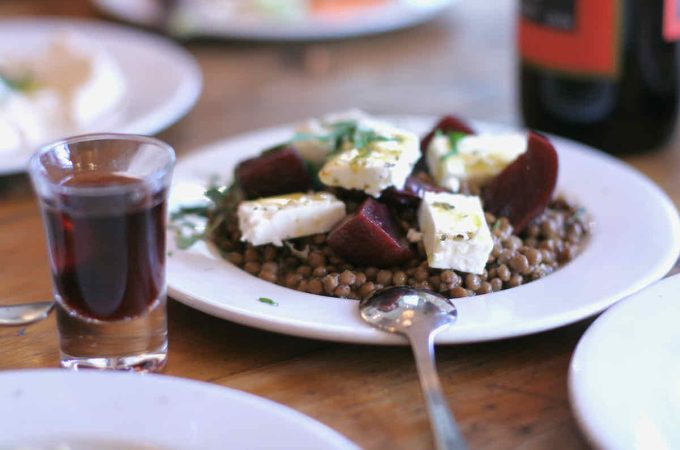 A white plate of puy lentil salad topped with large pieces of feta cheese and beetroot as served at The Real Greek restaurant at Westfield Stratford, East London. Adjacent is a shot glass of red sweet wine. A serving spoon, handle facing towards the foreground, lies face down on the plate. In the background of the vintage wooden table, a wine bottle with red label and a white plate with another side dish are shown in soft focus.