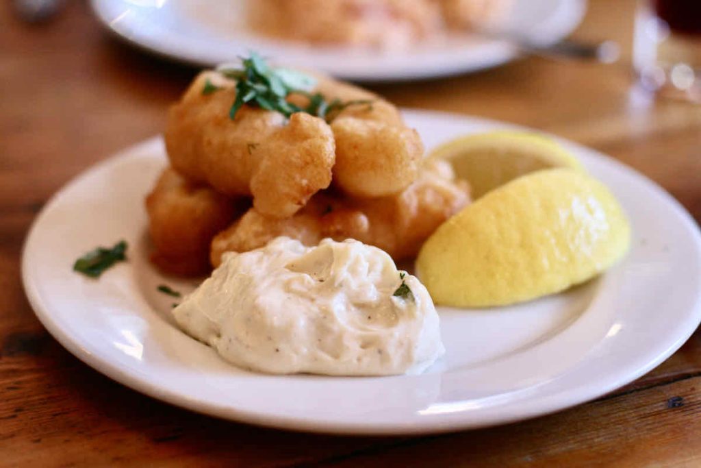 Salt cod fritters, scattered with green herbs, piled on a small plate with lemon wedge and lemon mayonnaise, as served at The Real Greek restaurant at Westfield Stratford, East London. On the vintage wooden table, another white plate of food lies in soft focus in the background. 