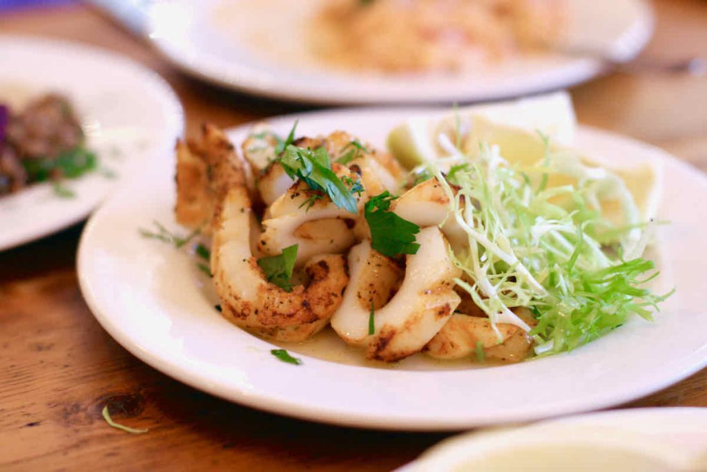 Slices of chargrilled calamari scattered with herbs and garnished with spiky lettuce salad leaves and lemon wedge, as served at The Real Greek restaurant at Westfield Stratford, East London. Two further white plates of food lie in soft focus behind. 